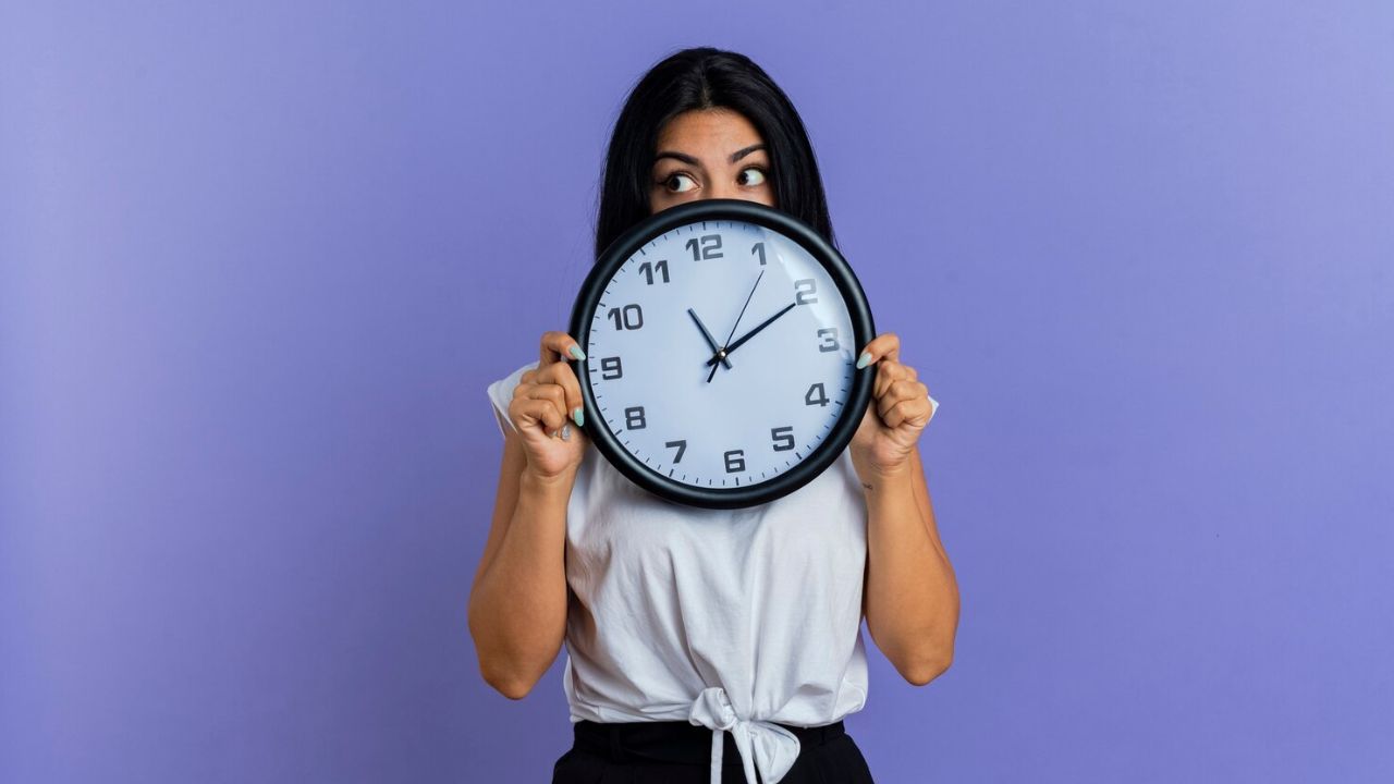 Joven de cabello largo cubriéndose la boca y la nariz con un reloj de manecillas que marca las 11:10 horas.