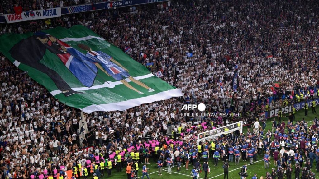 La hinchada del París muestra una pancarta de Luis Enrique y Xana clavando una bandera del PSG en un campo de futbol.